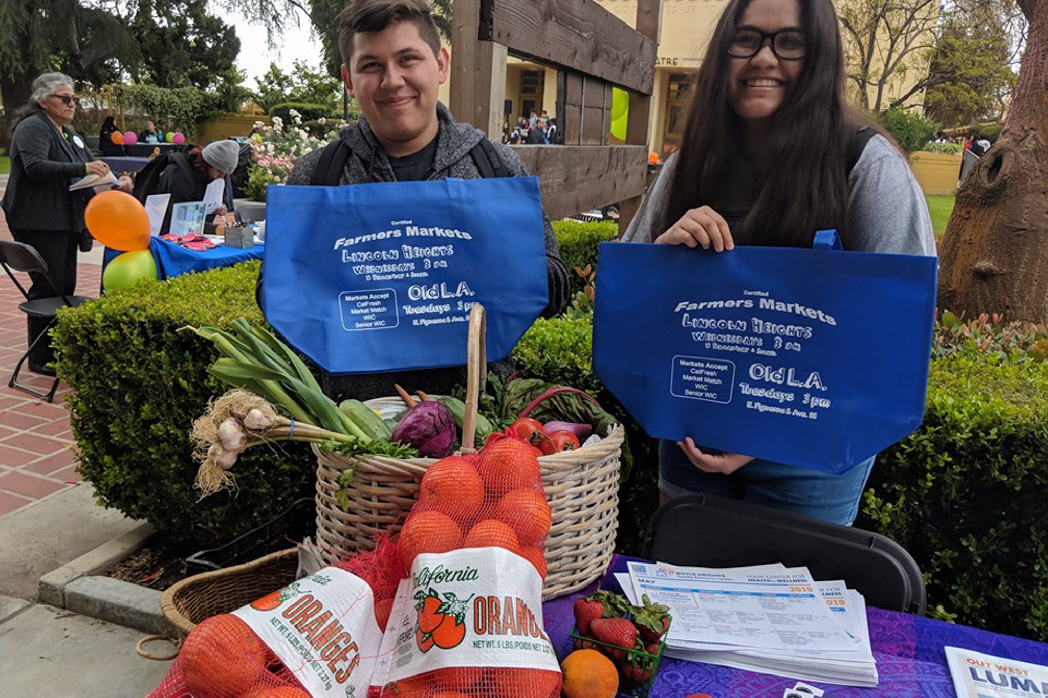 Community members holding Lincoln Heights Farmers Market tote bags with fresh produce and oranges